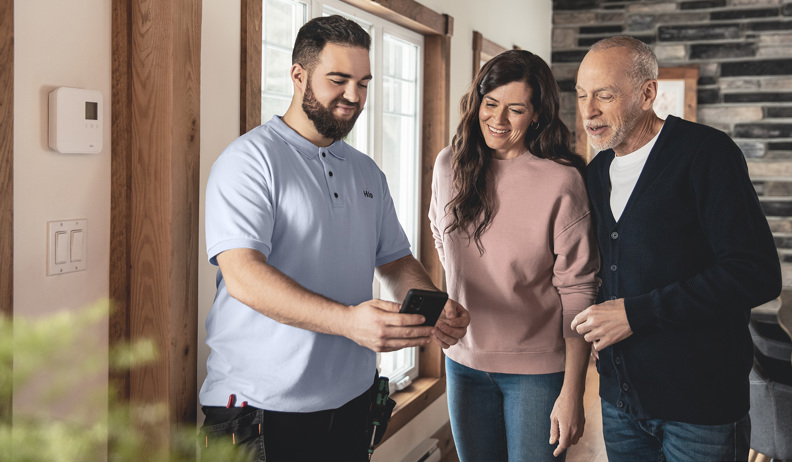 A Hilo installer with new Hilo customers look at a phone in a house with wood paneling and large windows; a smart thermostat is visible on the wall.