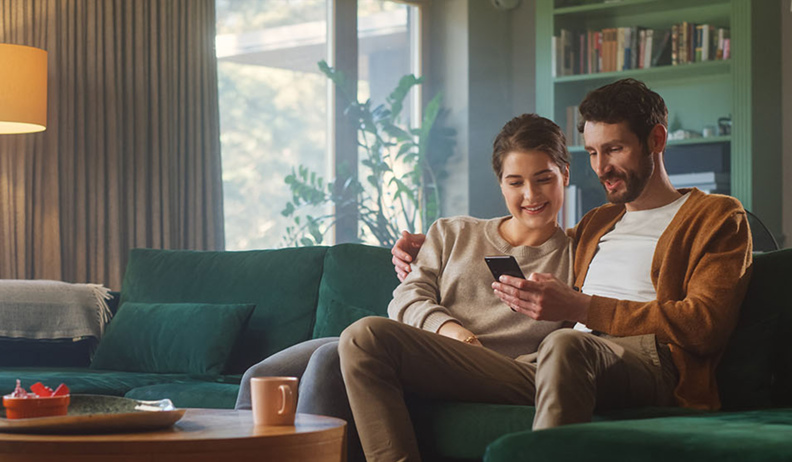 A smiling couple sitting on a green sofa look at a smartphone in a cozy living room with a lamp, shelves, and a coffee table.