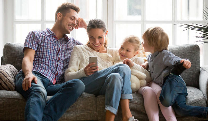 Two parents and two young children sitting on a sofa are looking at a smartphone in a bright room with large windows in the background.