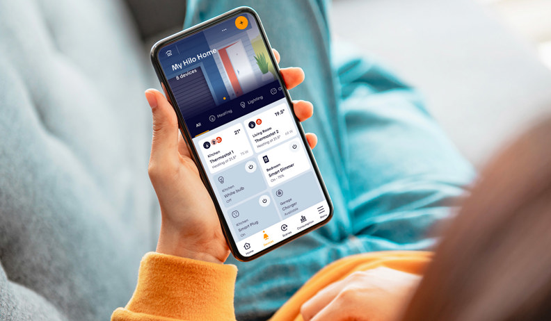 Person holding a smartphone showing the Hilo app with controls for lights, thermostats, and other connected devices.