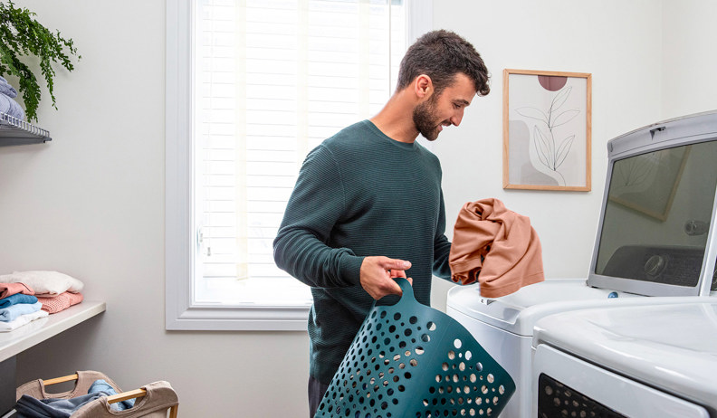 A man in a laundry room puts clothes in a washing machine, with a green basket, shelves, a window with blinds, and a frame on the wall.