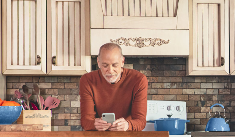 Person leaning on a kitchen counter looking at a smartphone; kitchen with wooden cabinets, fruit bowl, utensils, and blue pots and pans.